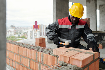 Experienced builder in workwear flattening surface of brick layer with special hammer while...