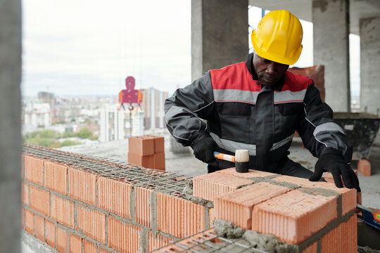 Experienced builder in workwear flattening surface of brick layer with special hammer while standing inside unfinished construction