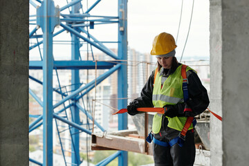 Young woman in protective helmet and reflective vest fastening safety belt on her waist while going...