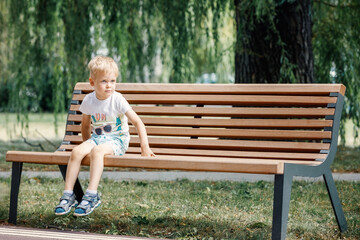 A cute boy is sitting on a bench in the park under a big green tree and resting.