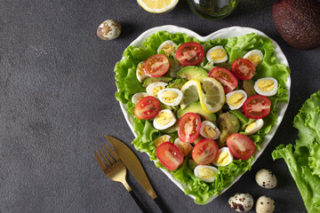 Salad of avocado, cherry tomatoes and quail eggs, served on green lettuce leaves in a heart-shaped plate, top view
