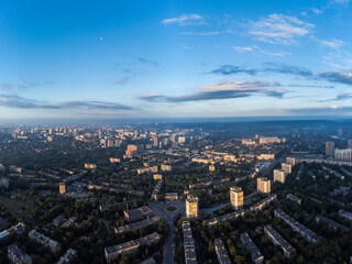 Naklejka na ściany i meble Early morning cityscape panorama view in summer green city, residential district with moon in blue sky. Aerial cityscape above buildings and streets, Pavlovo Pole, Kharkiv Ukraine