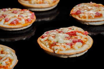 Many mini pizzas with ham, cheese baking on tray in electric oven, black background - close up view. Italian cuisine, homemade bakery, fast food, cooking concept