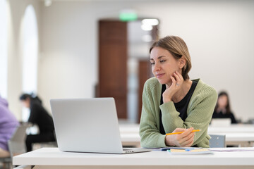 Middle-aged woman using laptop computer for e-learning, mature female student sitting in light...