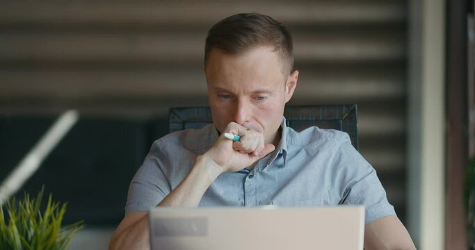 Concentrated man sits at desktop focused on a laptop screen. Turns the pen in his hand. Looks thoughtfully to the side