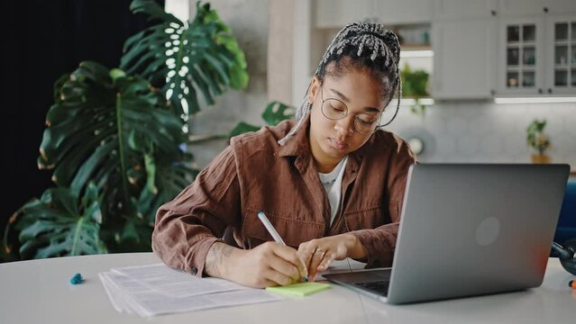 Young focused african american woman employee writing note and marking documents, working online on laptop, zoom out
