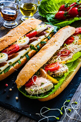 Long sandwiches with sausage, ham, chicken breast, boiled eggs, lettuce, arugula yellow cheese, tomato and dressing on wooden table
