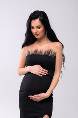Cropped of allure pregnant woman with evening makeup and circle eaarings, wearing black dress with open neckline, touching belly and looking down while standing against white bakground