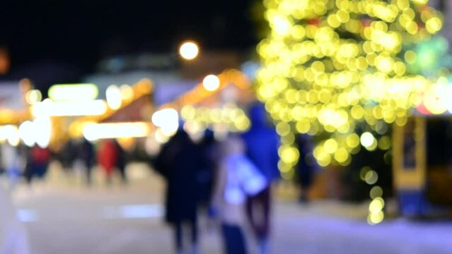 Blurred background. Christmas tree, decorated glowing lights, building, blurry silhouettes of people walking city square at winter night. Beautiful New Year and Christmas holiday blurred background