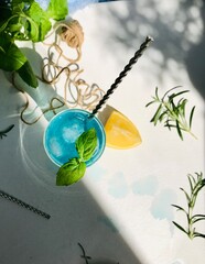Blue cold drink in a glass with ice cubes on white desk with blue background. Also used mint leaves and rosemary, rope, spoon  with natual sunlight from the open window