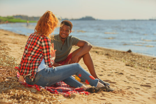Side view of smiling Black man listening to red-haired White woman while couple or friends talking sitting on blankets on sea beach