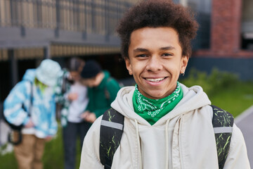 Portrait of African teenage boy with backpack behind his back smiling at camera standing outside of school building