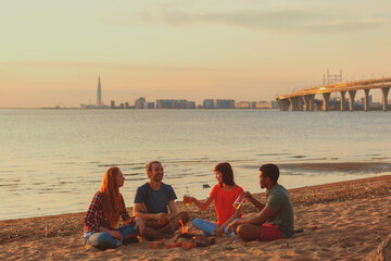 Group of four cheerful diverse young friends sitting around campfire on beach, drinking beer and...