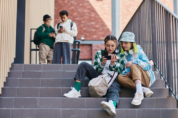 Obraz na płótnie Canvas Two teenage girls sitting on stairs outdoors and watching video on mobile phone with young boys in background