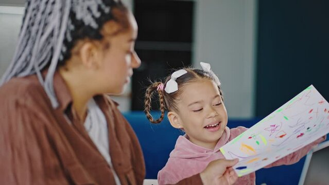 Cute little african american girl showing colorful picture to busy mother, woman working on laptop at home