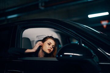horizontal photo from the side, at night, of a woman sitting in a black car and looking out of the window fixing her hair while looking in the side view mirror