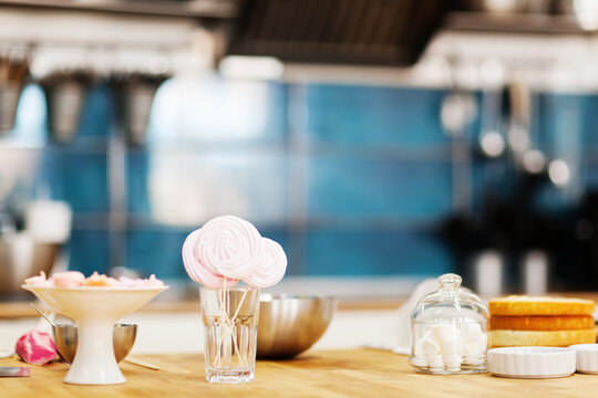 Meringue pops, marshmallows, sponge layers and bowls on table in bakery or pastry shop kitchen, background with confectionery ingredients and copy space