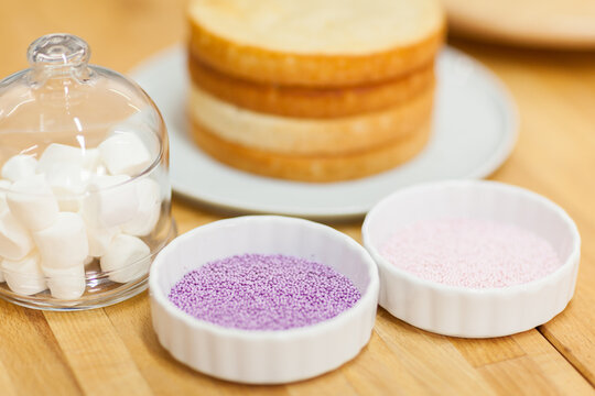 Sugar bead for cake decoration in saucers, marshmallows in glass jar and sponge layers for cake on table. Background with pastry shop ingredients