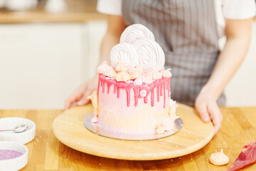 Freshly baked drip cake decorated with meringues placed on rotating stand in bakery kitchen. Pastry chef showing ready cake