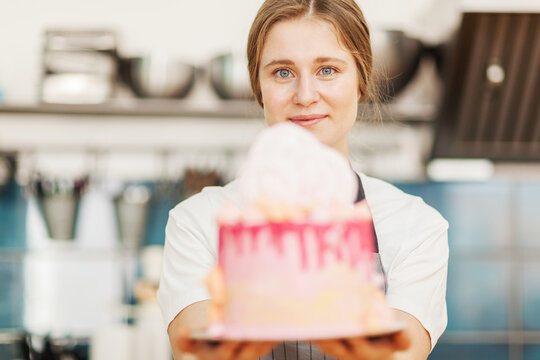 Prettyl female pastry chef looking at camera while demonstrating defocused handmade drip cake standing in restaurant kitchen