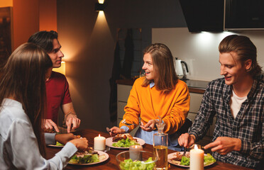 Group of four young friends enjoying roasted chicken and friendly talk sitting at table during cosy home dinner