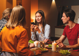 Group of four young friends having dinner together in domestic kitchen. Young woman reading text message or news on cell phone to friends