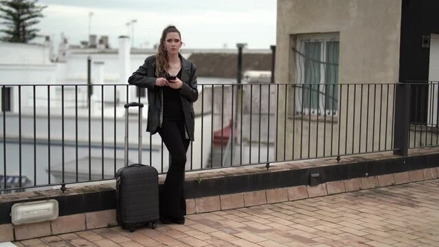 Chica delgada joven llegando a hotel con maleta de ruedas y smartphone