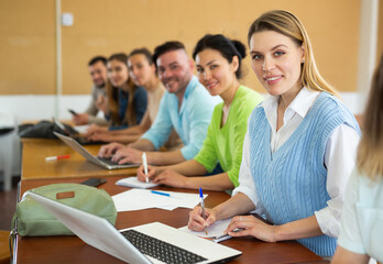 Obraz premium Woman university student sitting at table in classroom and exercising, writing in copybook and using laptop. University students sitting in a row.