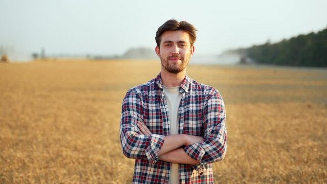 Portrait of happy farmer standing in ripe wheat field with arms crossing on chest. Proud agronomist looking at camera on a farmland. Rich harvest of cultivated cereal hybrid crop. Harvesting season.