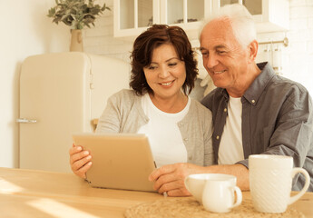 An elderly couple sits in the kitchen and uses a tablet, watches the news, enjoys spending time together.