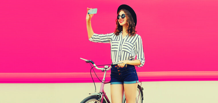 Beautiful happy smiling young woman taking selfie by smartphone with bicycle wearing shirt, shorts, black round hat on pink background