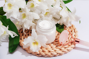 Obraz na płótnie Canvas Cream jar and rose quartz facial roller with big white flowers. Beauty, spa and wellness concept. Top view