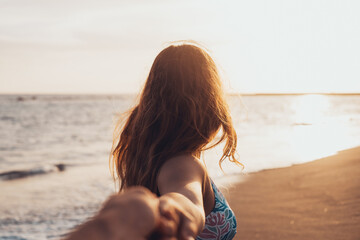 Portrait and close up of young woman holding hand of boyfriend walking on the beach smiling and having fun together. Man following girlfriend to the water of the sea enjoying.