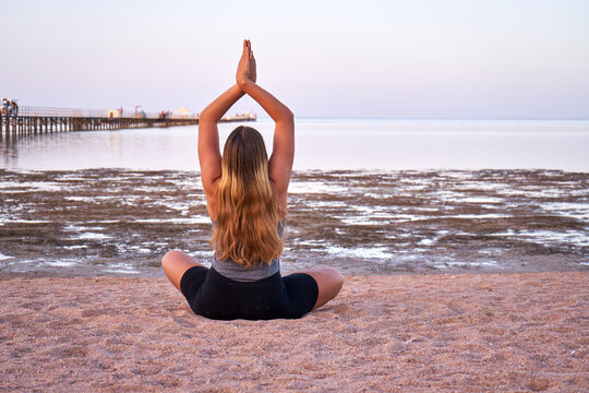 Young long hair woman practicing yoga on the beach at sunset. Caucasian girl with long blond hair doing yoga exercises on a sandy beach at sunrise.   