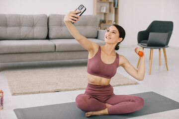 Fototapeta na wymiar Attractive young woman taking selfie showing biceps with dumbbell at home.