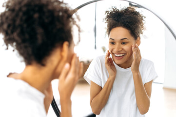 Satisfied beautiful African-American girl with curly hair in a white t-shirt, stands in front of a mirror, examines her face, rejoices at the healthy state of her facial skin, smiling happily