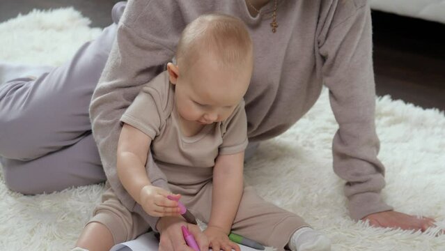 Mom, baby play together and draw at home in room. Mother teaches baby son to draw with colored markers on paper sheet. Careful woman and child sit on white fluffy rug in room at home close view.