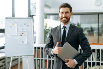Fototapeta premium Elegant positive Caucasian businessman, company ceo, auditor, stands in a modern office, in a formal business suit, with a laptop in his hands, looks at the camera, smiles, shows thumbs up gesture