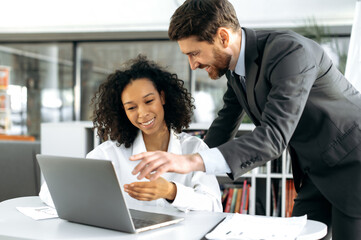 Fototapeta premium Positive smart multi ethnic colleagues, caucasian man and african american girl, web developers, in modern office working on laptop, writing an algorithm for a mobile application, discussing details