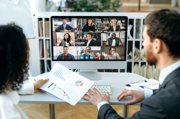 Two business multiracial partners, a man and a woman, are sitting in a modern office, in front of a computer screen, conducting online brainstorm for an international group of company employees