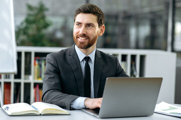 Handsome positive Caucasian businessman, company manager, financial consultant, sits at a workplace in a modern office, in formal suit, works at a laptop, looks to the side, analyzes work tasks, smile