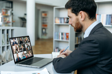 Fototapeta na wymiar Distant video conference. Focused Caucasian male, office worker, sit at a desktop in front of a laptop screen, listens an online business briefing with international colleagues by laptop, take notes