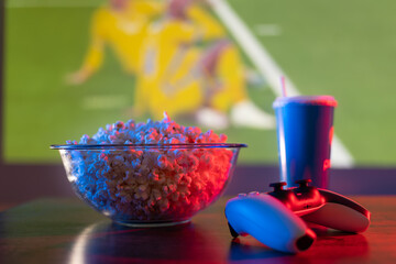 Video games. Popcorn, drink and joystick on the background of a large TV screen with a sports game. Football, basketball, volleyball, American football. Home comfort, attributes of a sports fan. © Anton