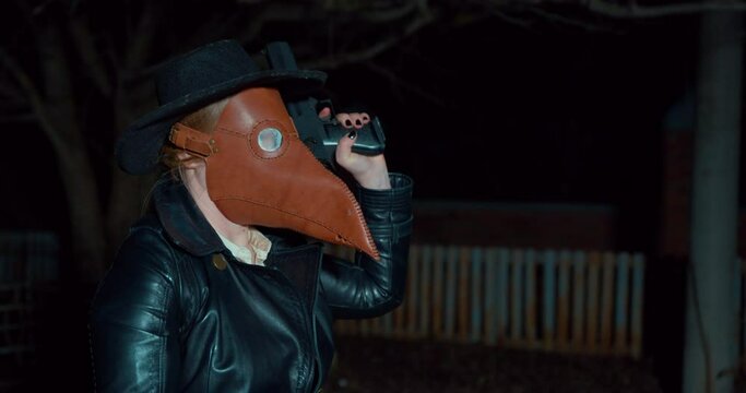 Person wearing hat, leather coat and plague doctor mask aims a gun at victim or enemy, and then fires another weapon, side view. Criminal hid face to commit a robbery