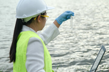 Female environmentalist wearing gloves takes samples of river water to examine contaminants in natural waters and record data on a laptop. Water and ecology concept.
