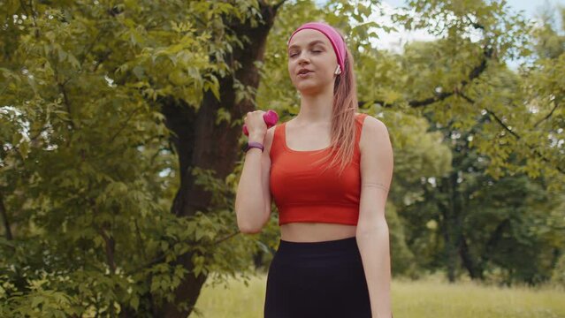 Athletic sporty fit girl in sportswear doing dumbbells workout in park performing bicep curls to build arm muscle and strength. Young woman enjoying exercising outdoors. Active sportswoman. Motivation
