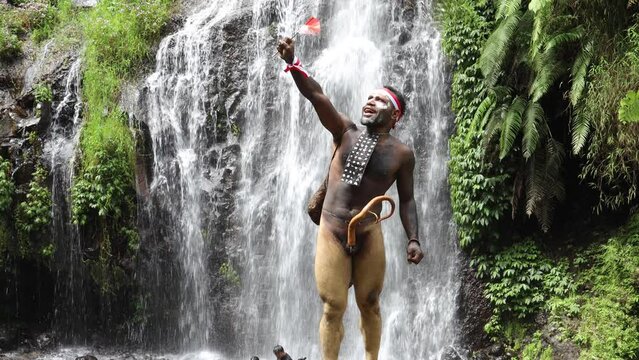 Papua man wearing traditional clothes of Dani tribe, red-white headband and bangle is holding little Indonesia flag and celebrating Indonesia independence day against waterfall background