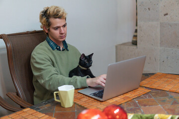Blond boy using his computer in the dining room of his house next to his cute cat