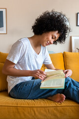 Young African American woman reading a book sitting on yellow couch. Female relaxing at home....