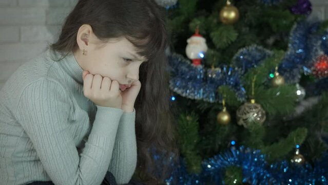 Child sad mood in Christmas decorations. A girl with sad mood pas her holiday time by the Christmas tree.